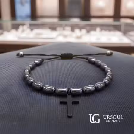 Pulsera hematita ovalada negra con cruz negra colgante y cordón ajustable para hombre, Black Series Ursoul Germany, sobre superficie azul marino en joyería