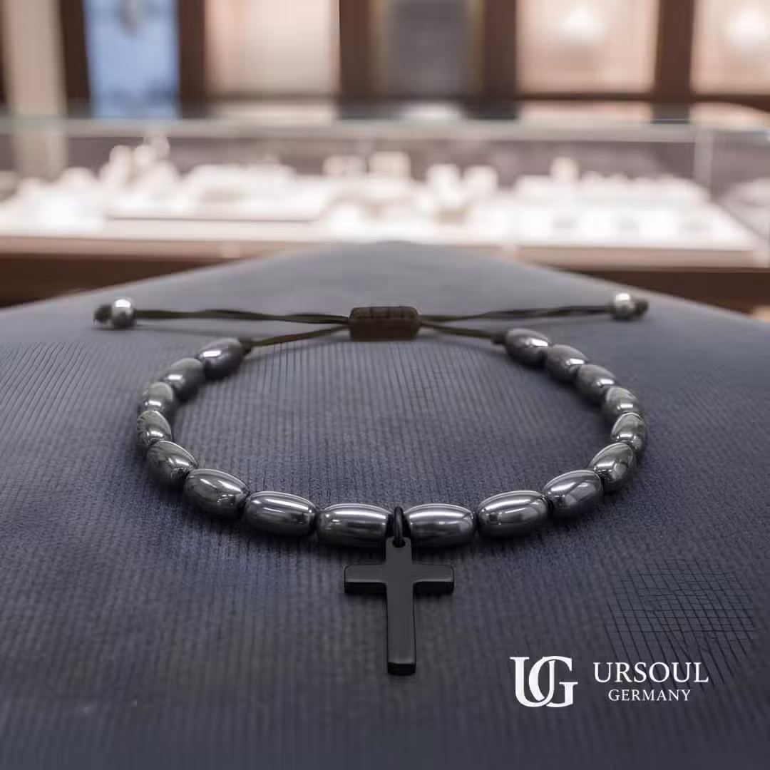 WhatsApp Image 2026-08-15 at 9.52.21 AM (5) Pulsera hematita ovalada negra con cruz negra colgante y cordón ajustable para hombre, Black Series Ursoul Germany, sobre superficie azul marino en joyería