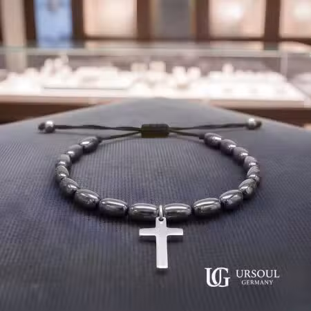Pulsera hematita ovalada negra con cruz plateada colgante y cordón ajustable para hombre, Silver Plated Ursoul Germany, sobre superficie azul marino en joyería