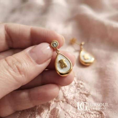 Aretes gota de nácar con osito dorado y cristal 2.5cm para mujer, Gold Color Ursoul Germany, en mano sobre tela rosa y fondo crema