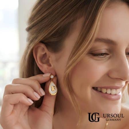 Aretes gota de nácar con osito dorado y cristal 2.5cm para mujer, Gold Color Ursoul Germany, en mano mientras mujer sonrie