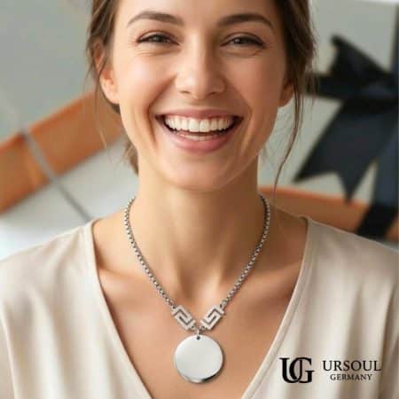 Collar medallón circular plateado con llave griega para mujer, Silver Plated Ursoul Germany, sobre pizarra gris con cajas de regalo y sobre escote en V con blusa beige