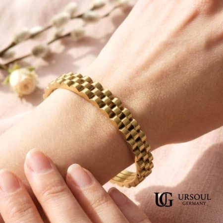 Brazalete rígido dorado diseño presidencial para mujer, Gold Color Ursoul Germany, sobre mármol blanco y sobre muñeca con fondo floral rosa